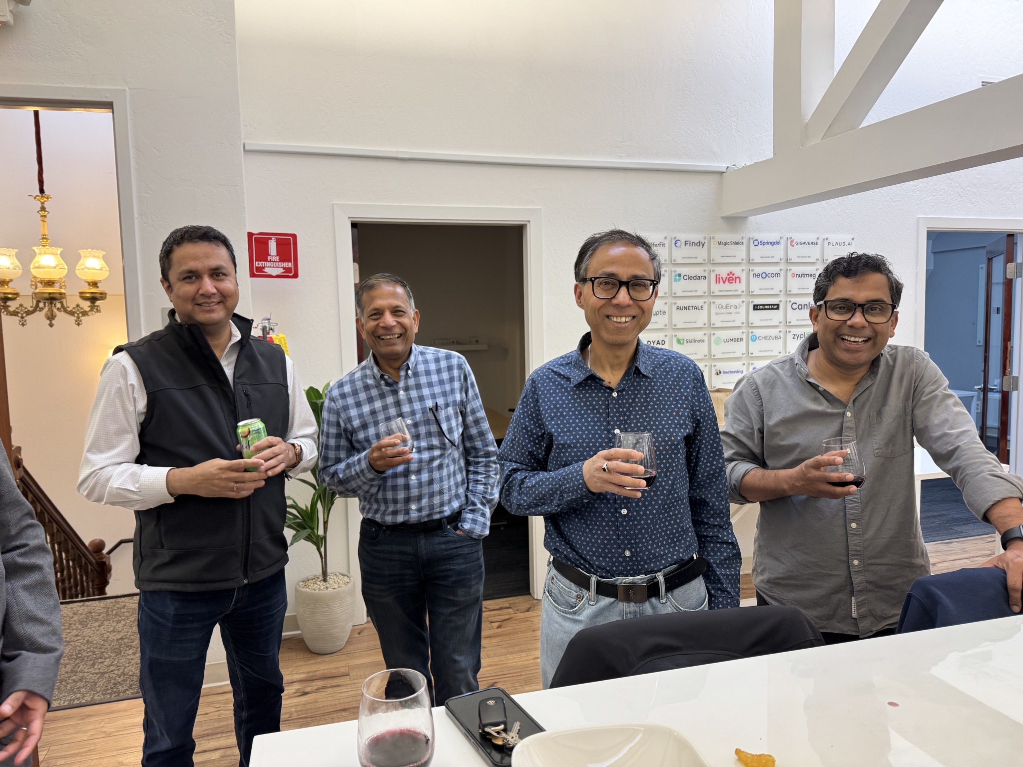 Four men stand together in a modern office space, holding drinks and smiling. One holds a can, three hold wine glasses.