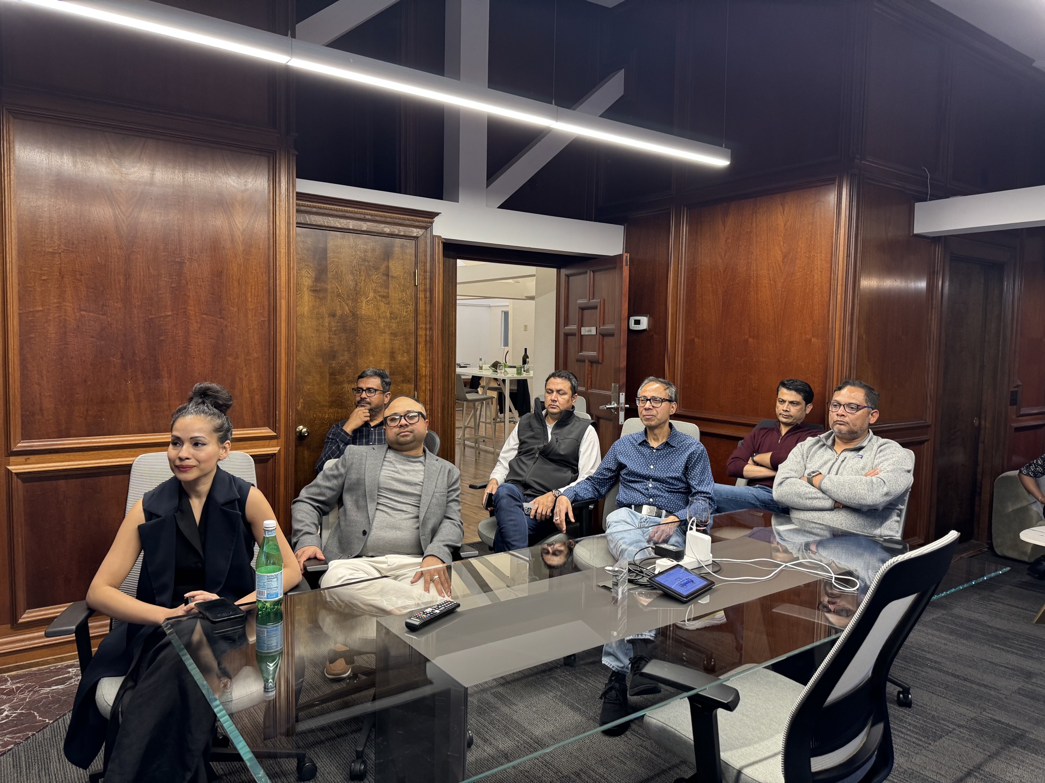 A diverse group of entrepreneurs sit around a glass table in a wood-paneled room, listening intently during a meeting.