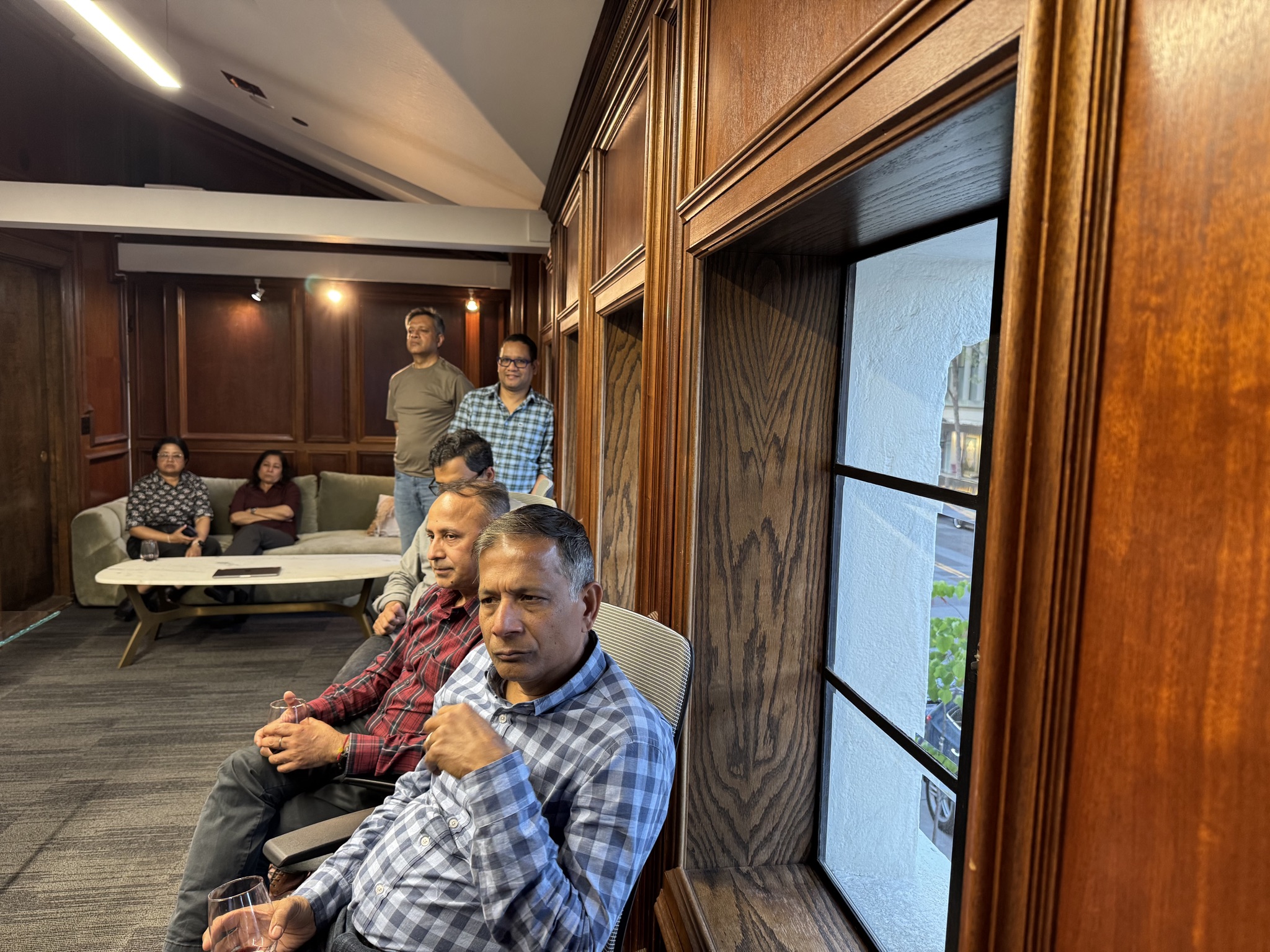 Several men and women are gathered in a room with wood paneling and large windows. Some are seated, holding drinks, while others stand.