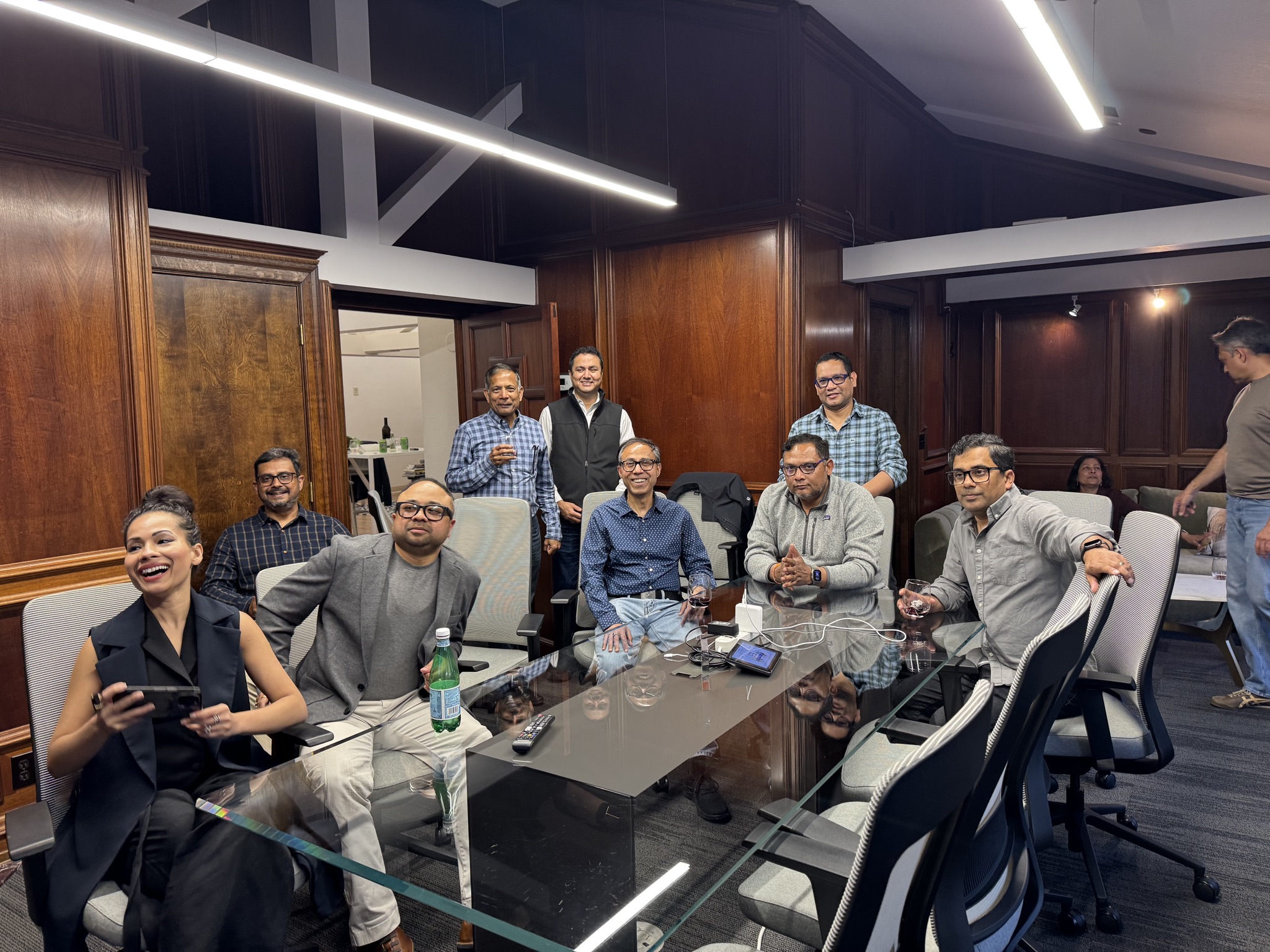 A group of entrepreneurs sits around a glass-topped conference table in a wood-paneled room, some holding drinks and looking towards the camera.