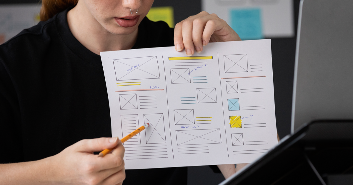 Osoba drží vytištěný wireframe webu a tužkou ukazuje na jednu z částí rozložení během diskuse o UX/UI designu.