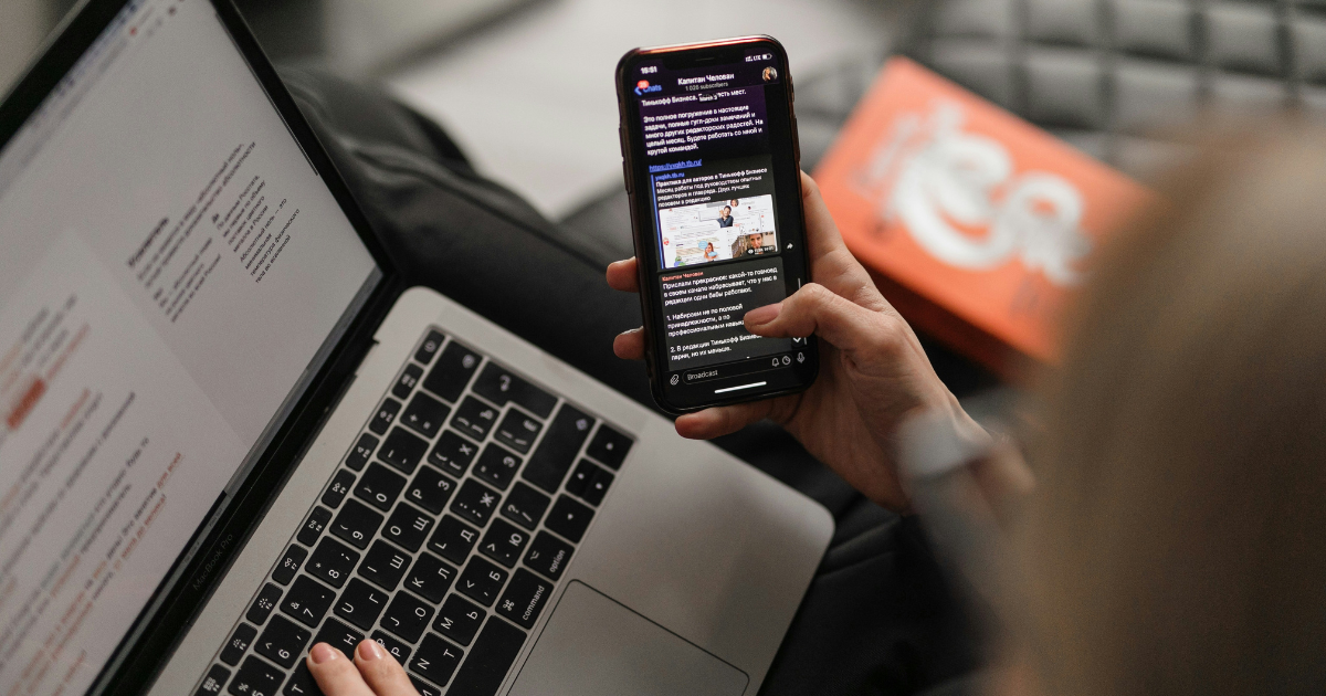 A person holding a smartphone while working on a laptop. 