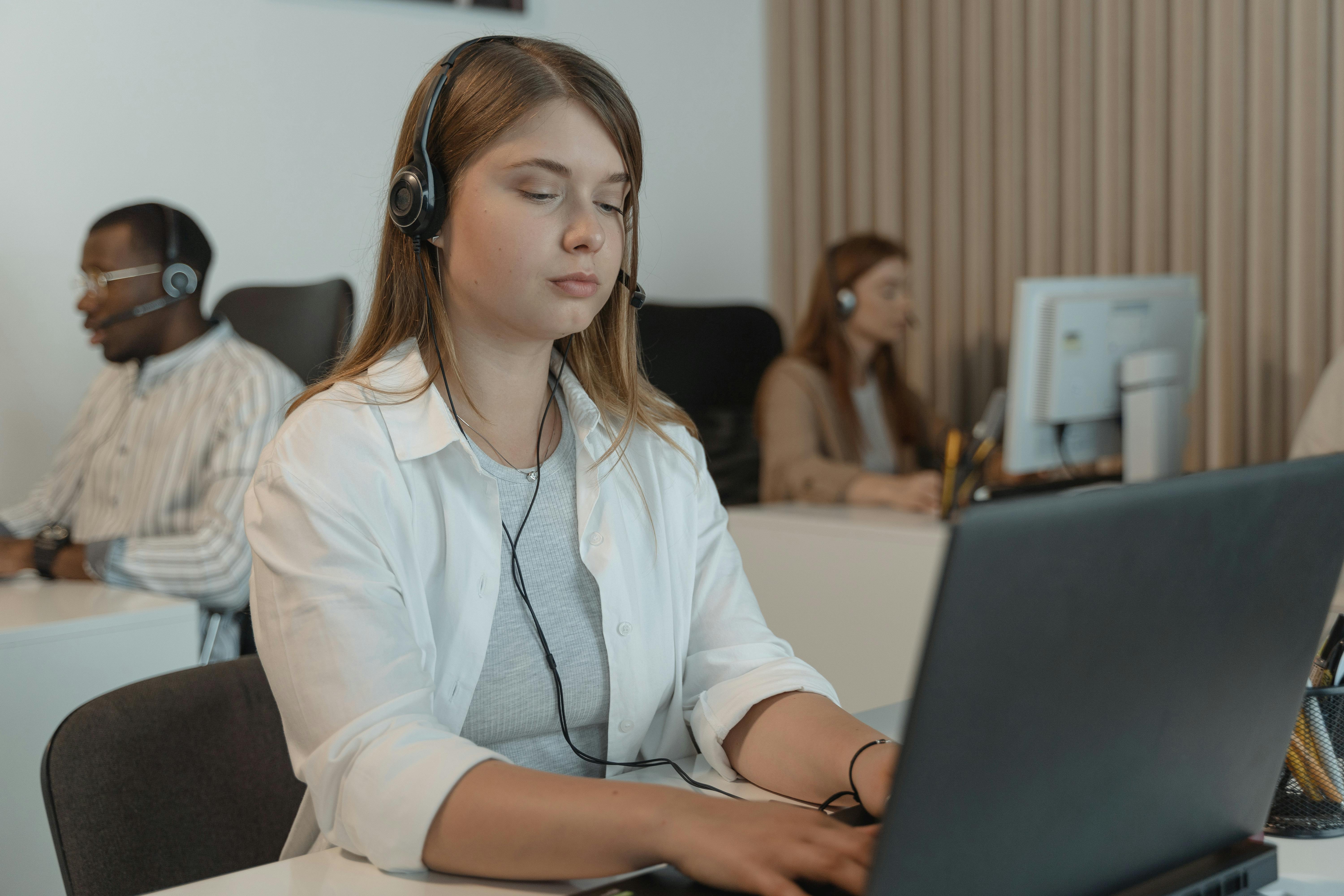 Young woman wearing a headset working on a laptop in an office with colleagues in the background also wearing headsets.