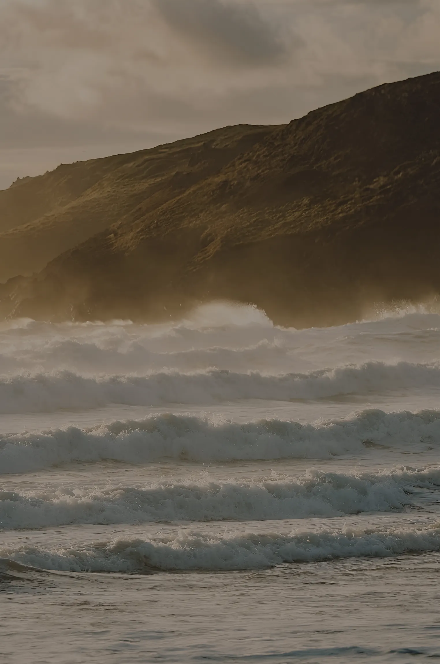 Ocean waves crashing near a misty rocky coastline at sunset under a cloudy sky.
