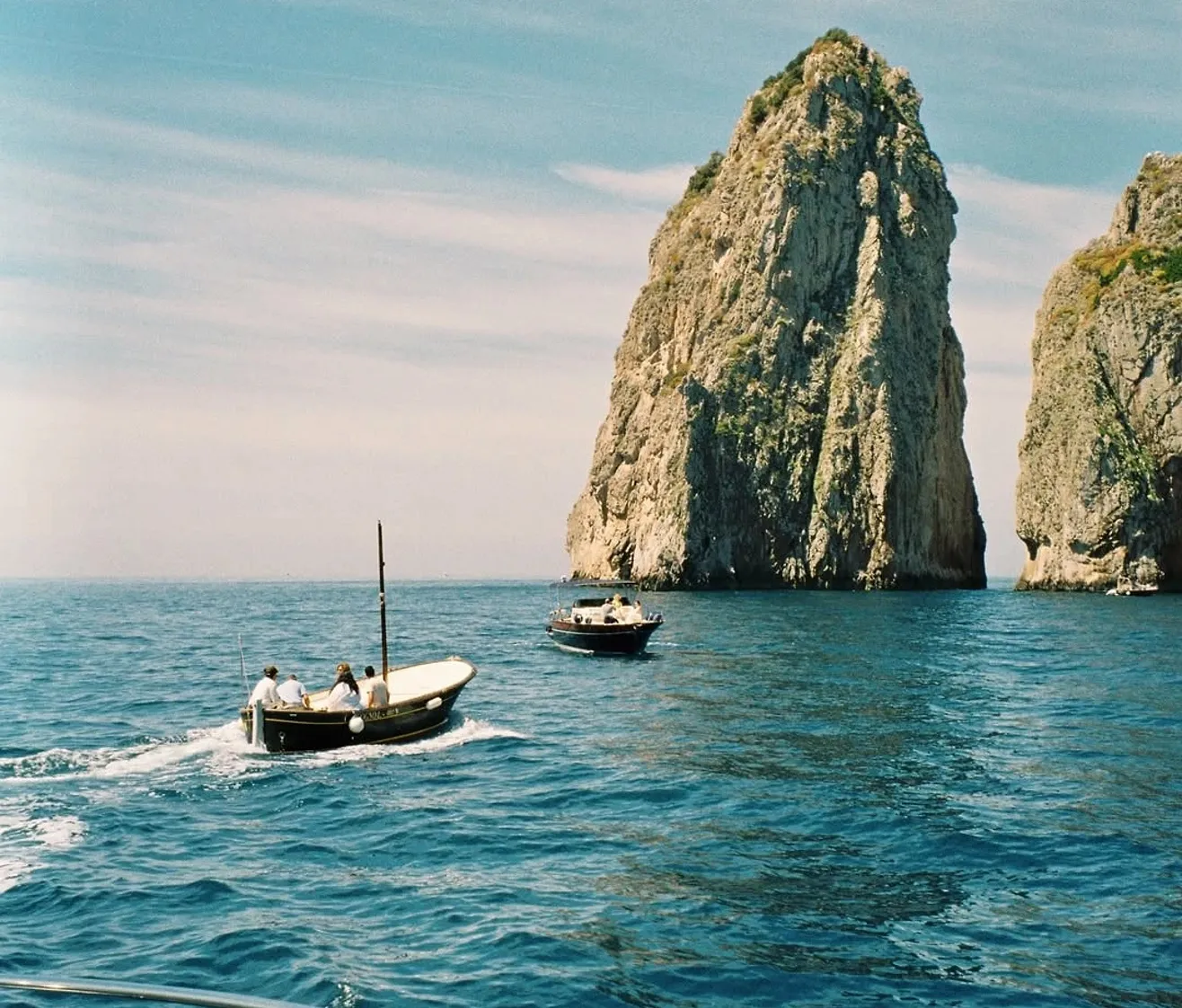 Two small boats with people sailing near large rocky sea cliffs on a clear day.