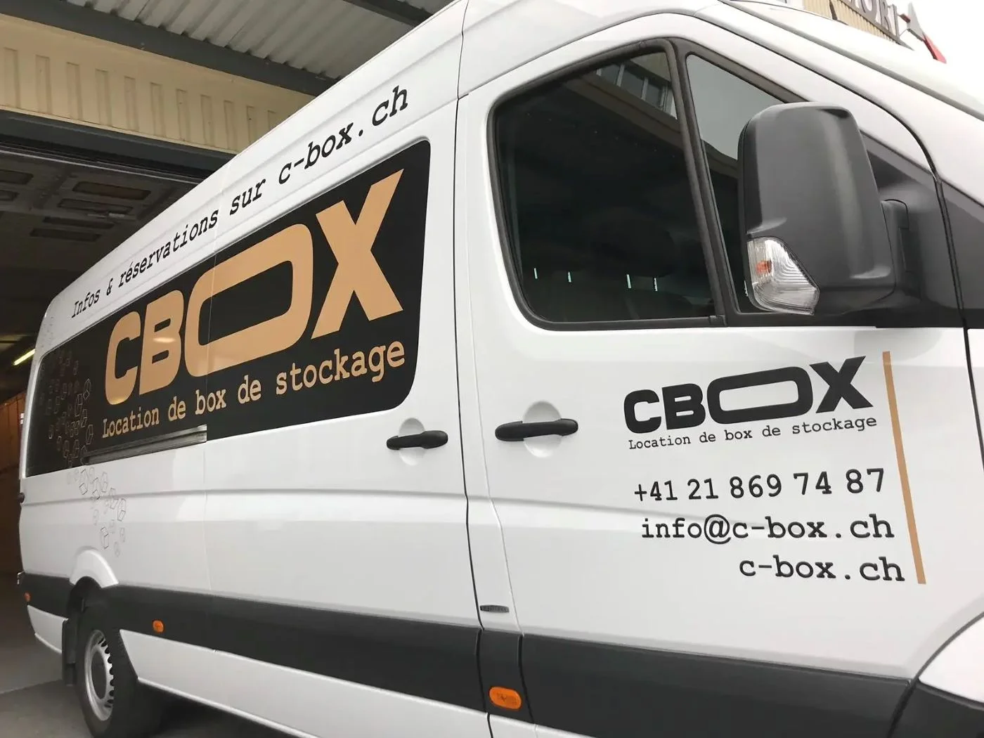 Camionnette de déménagement C-BOX blanche avec logo et informations de contact sur le côté, stationnée devant un bâtiment.