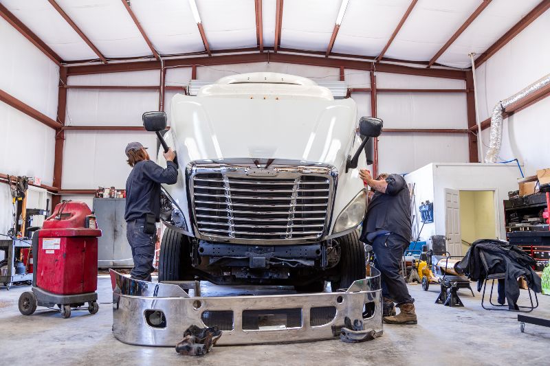 Two mechanics working on a white semi-truck inside a repair shop, front bumper removed, tools and equipment visible in the background.
