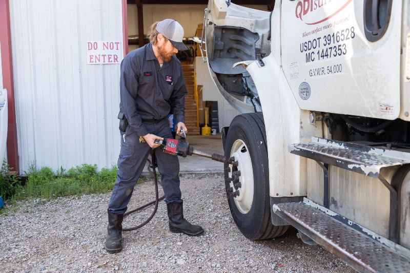 Heavy-duty truck mechanic using a pneumatic impact wrench to tighten lug nuts on a semi-truck wheel outside a repair shop.