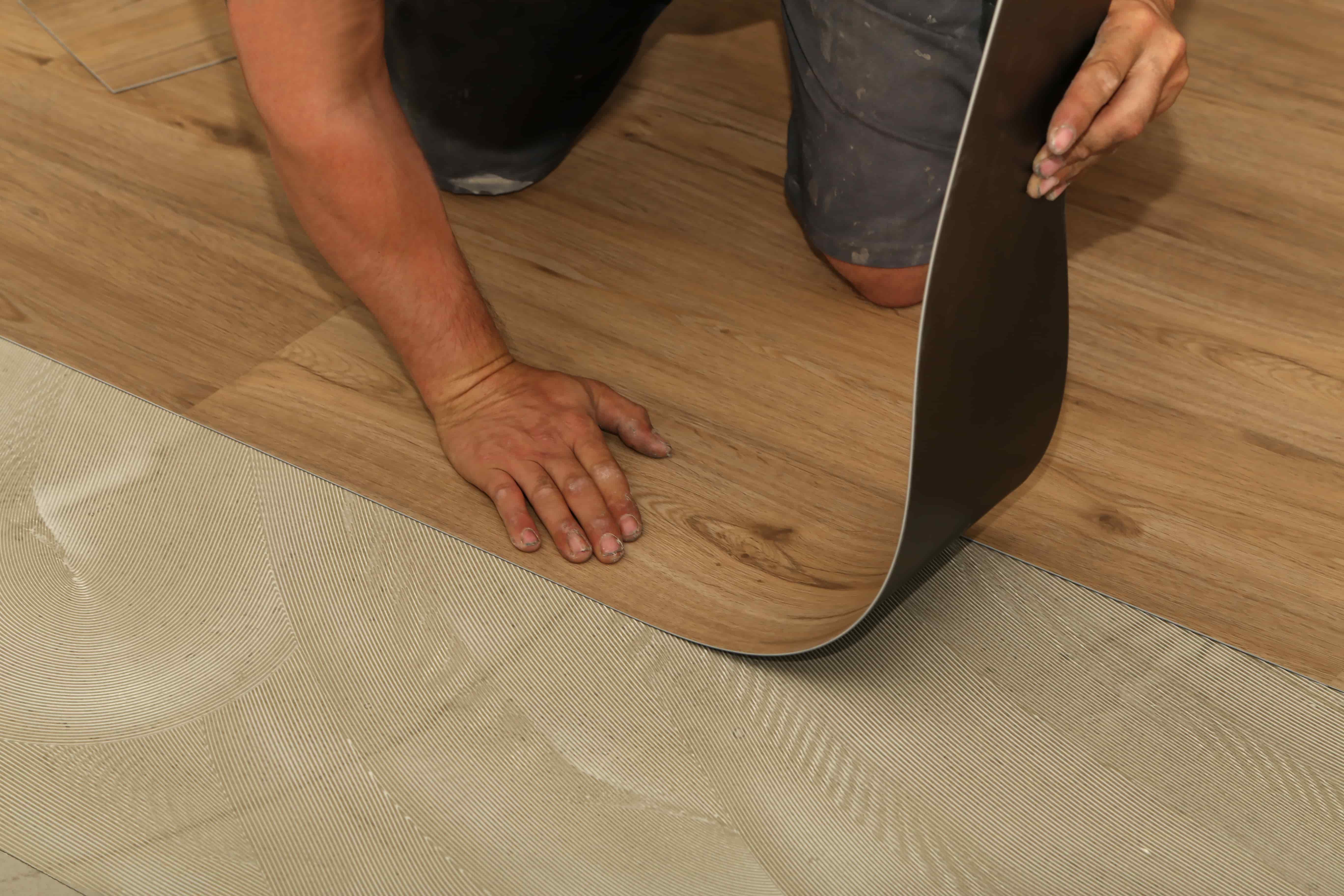 Vinyl plank boards are installed on a surface with wet glue. The installer is placing a board down gently by first fitting the end against the previous board.
