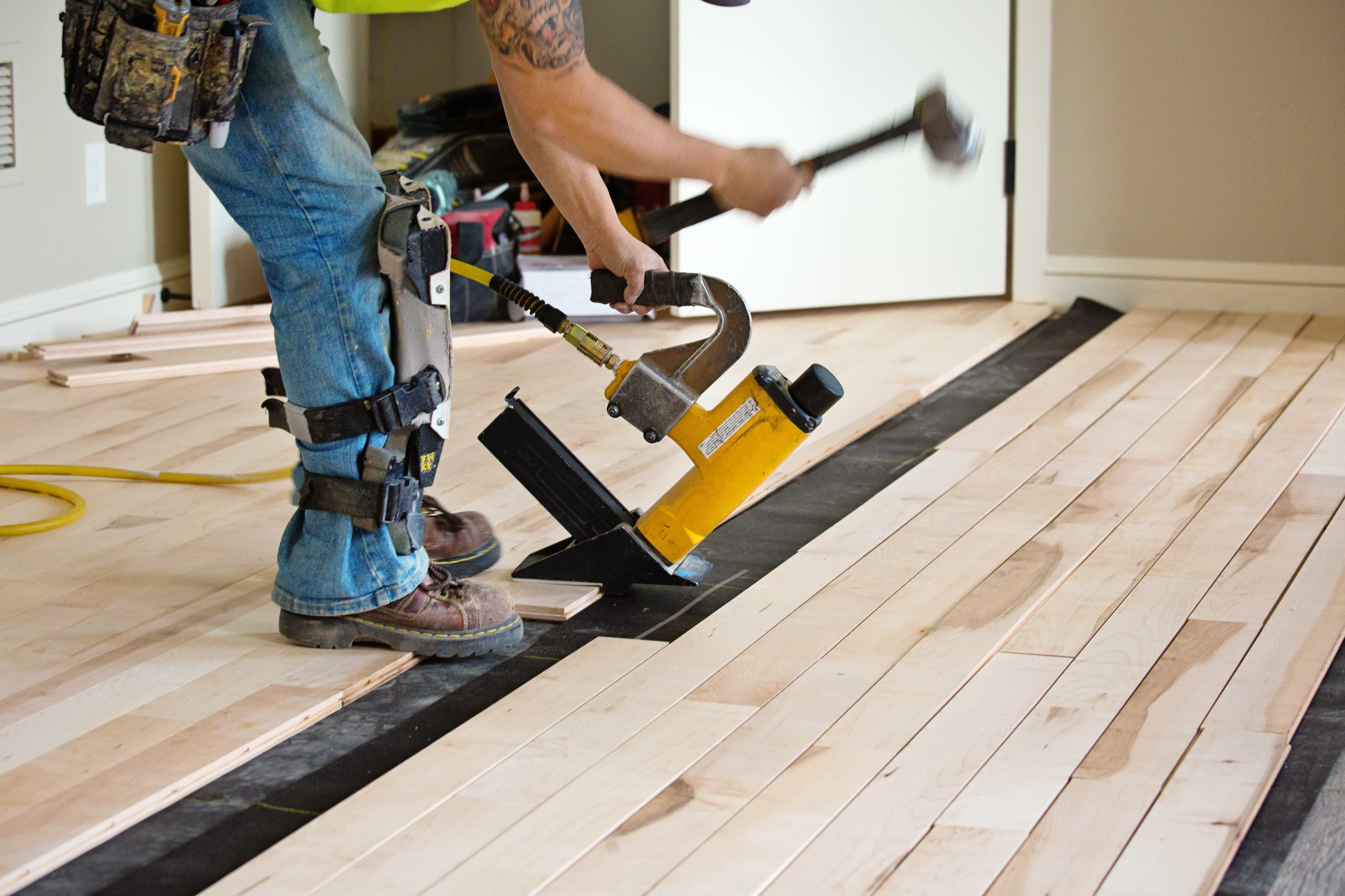Unfinished hardwood floor boards are being installed. The installer is dressed in work clothes and strikes the nailer with a mallet to nail a board down.
