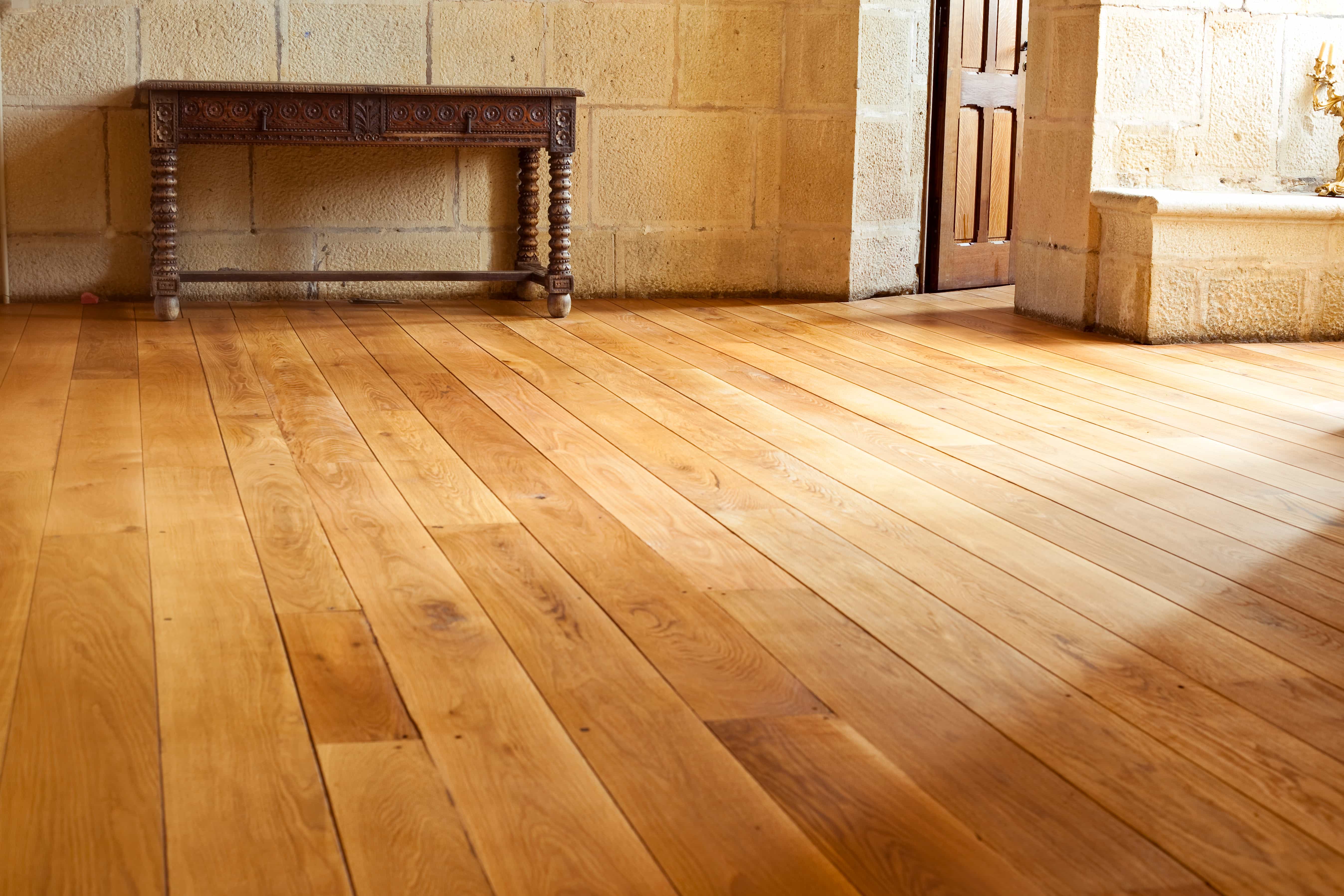 Rustic hardwood floors in the foreground with sun shining through from the right. The walls are light colored stone with an antique wooden bench against the back wall.