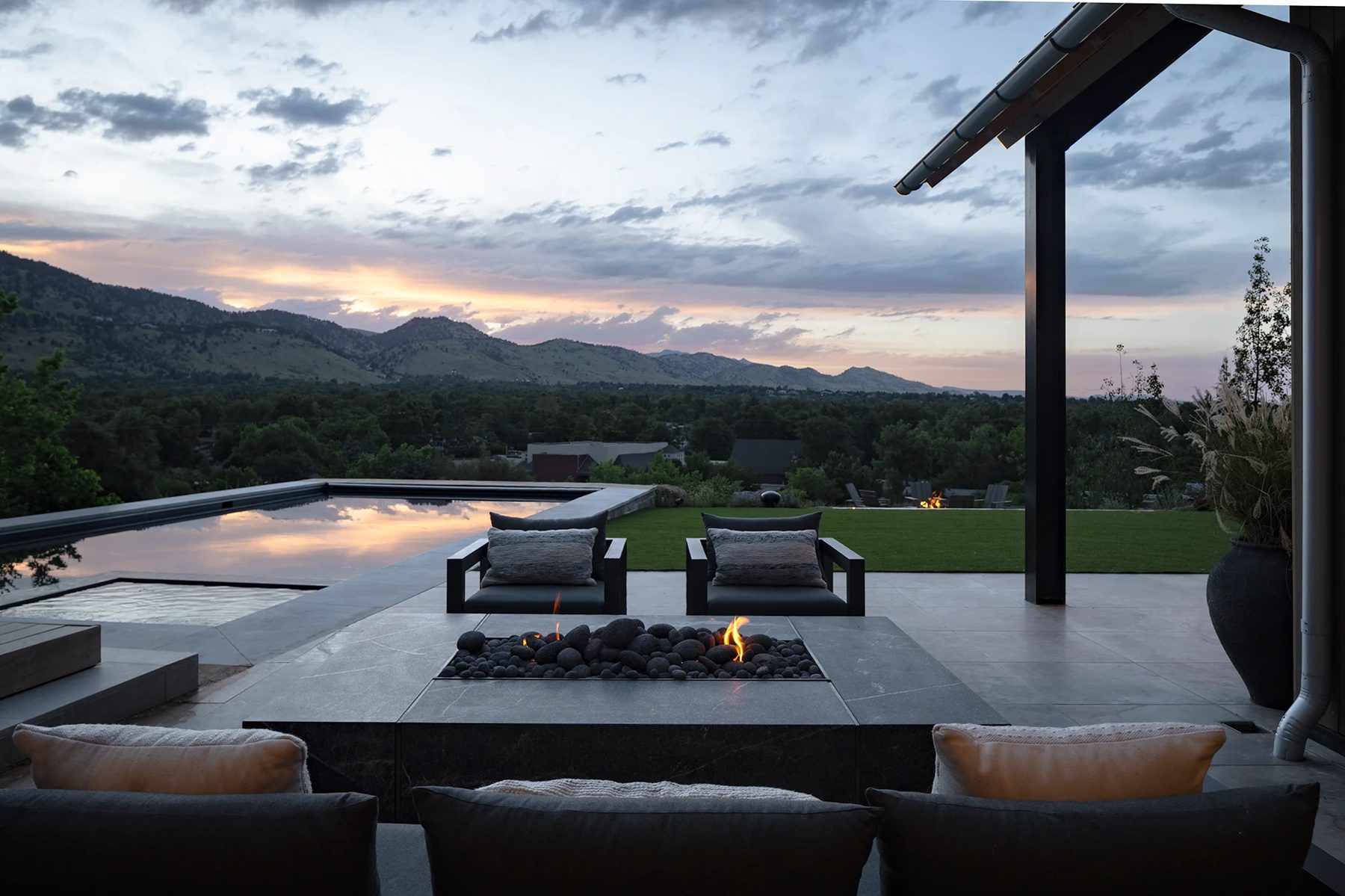 Twilight terrace with a long pool, a gas-style fire table, dark chairs and a lawn beyond
