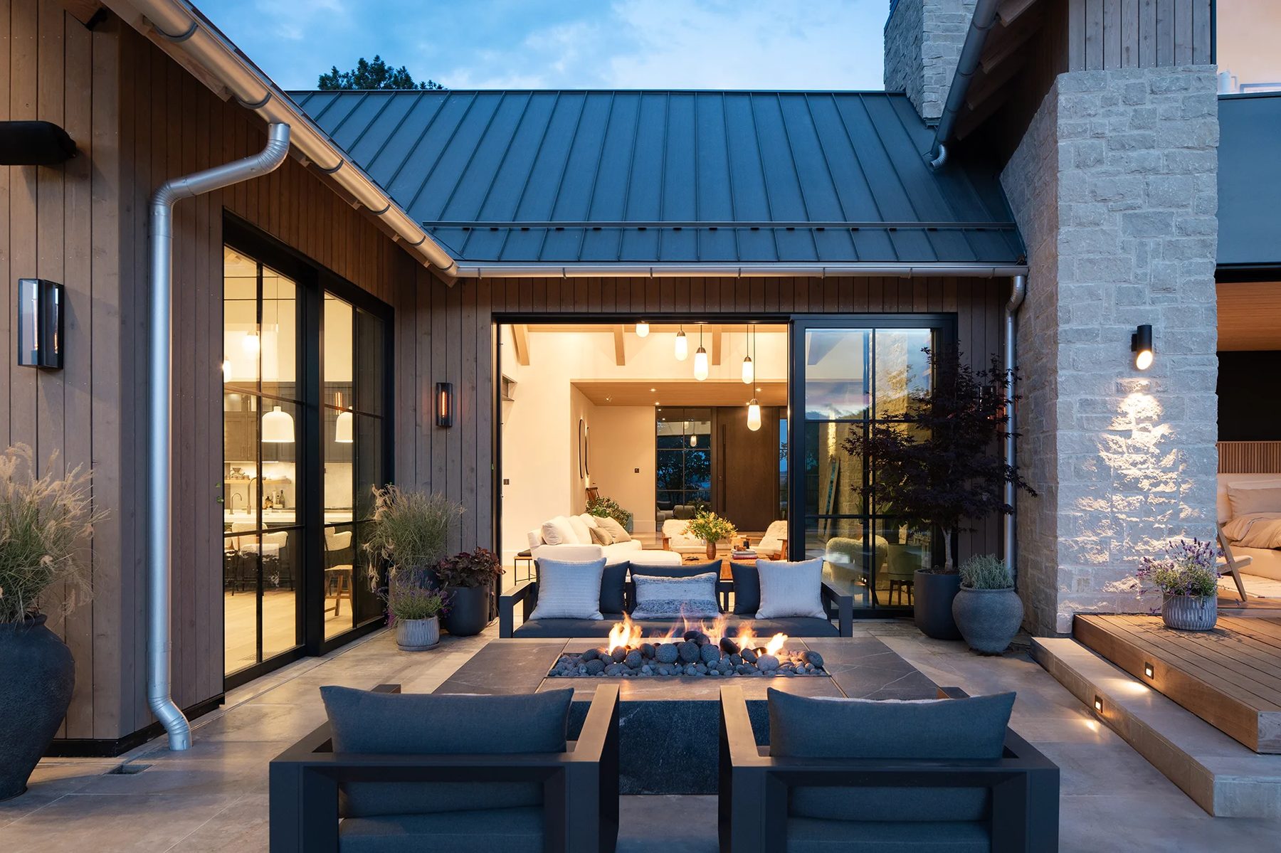 Dusk patio with a fire table, dark sofa and chairs, glass doors, stone column and lit wood steps