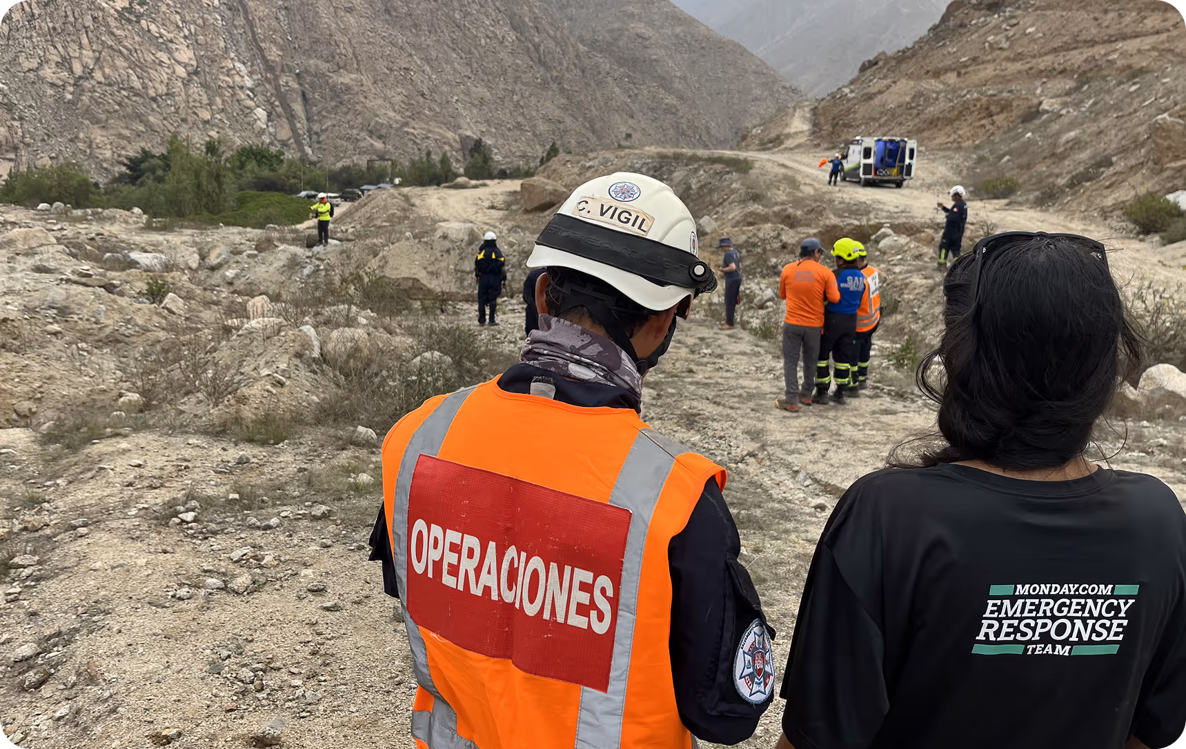 Emergency response team members in safety gear stand on rocky terrain near mountain rescue vehicle.