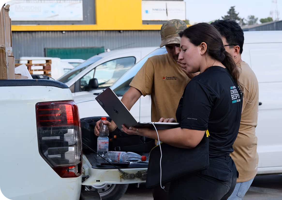 Three people stand near the back of a white pickup truck in an outdoor industrial area, looking at a laptop held by a woman wearing a black 'Emergency Response' shirt.