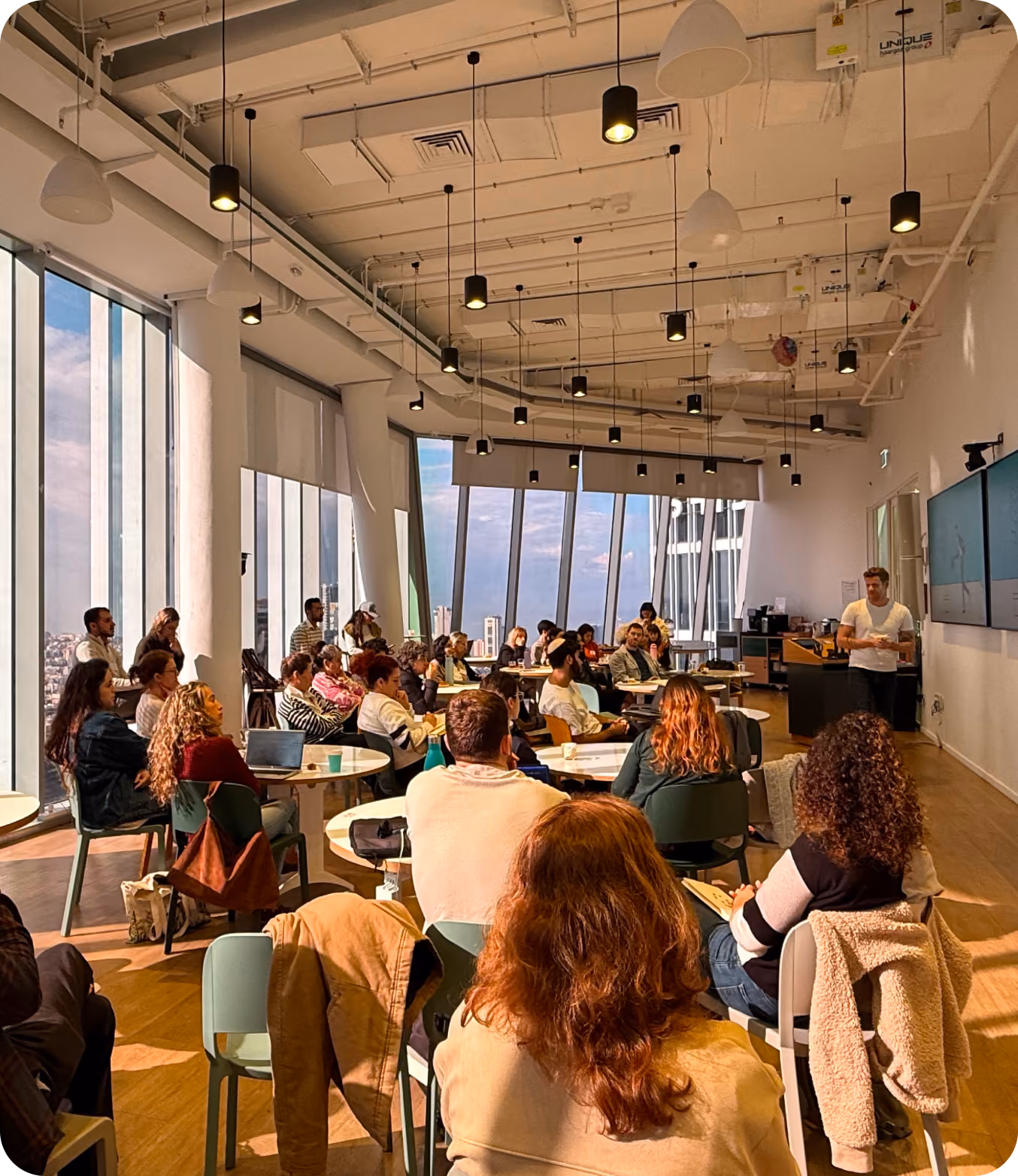 Non-profit ecosystem image: A modern conference room with large windows and ceiling lights, filled with an attentive audience seated at round tables listening to a presenter standing near two large screens on the wall.