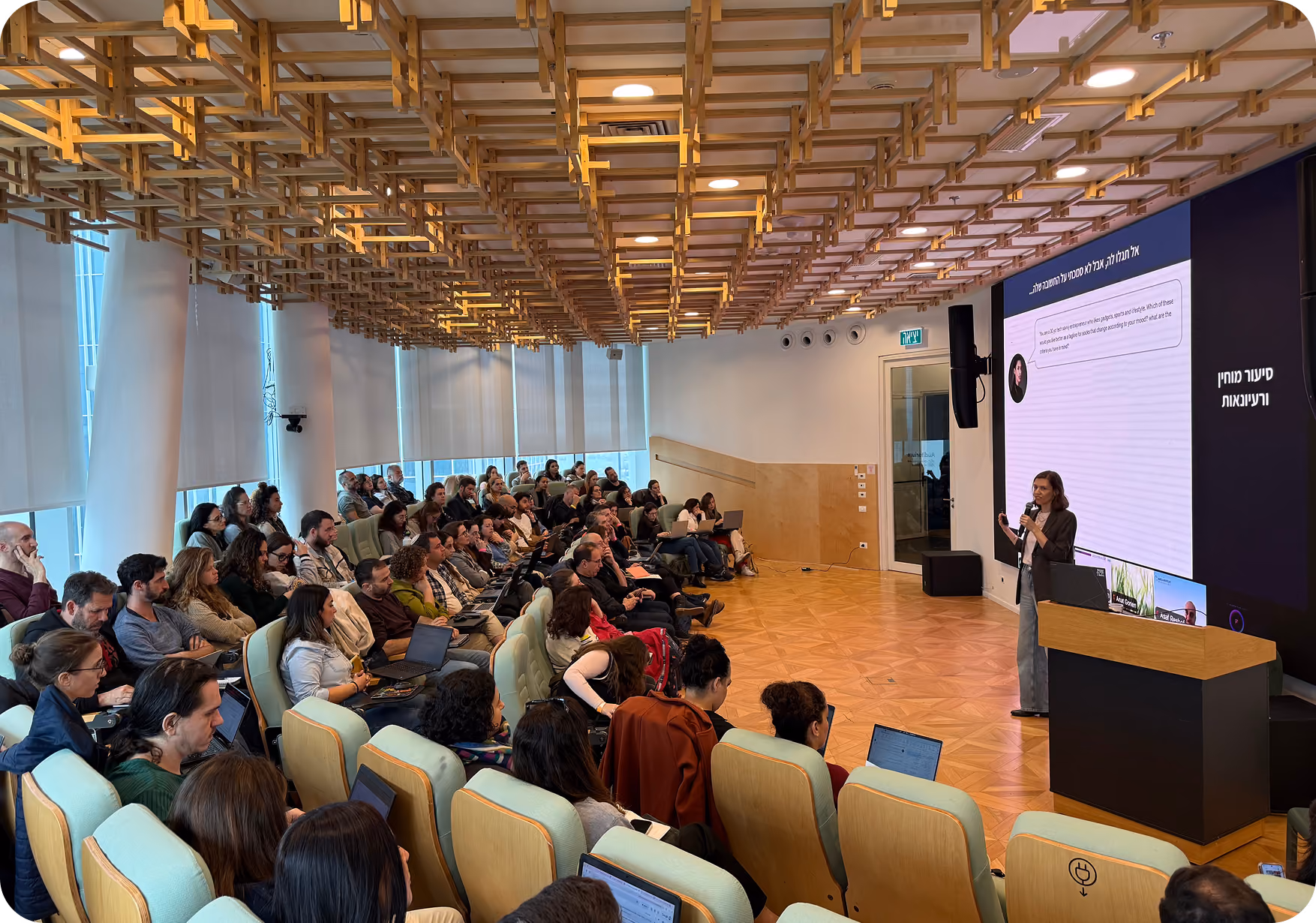 Access create action image: Speaker giving a presentation to an audience seated in a modern auditorium with wooden ceiling features and large windows, a projection screen displaying content.