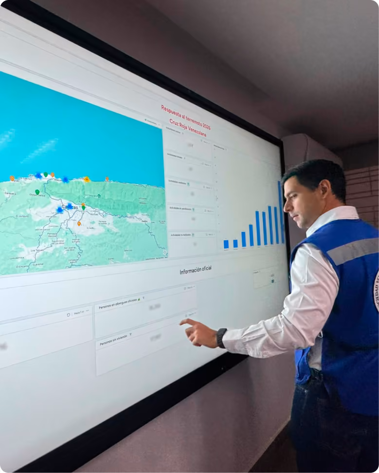 A man in a blue and white vest interacts with a large wall-mounted touchscreen displaying a map of Venezuela with colored markers and a bar graph, showing earthquake response data labeled 'Respuesta al Sismo 2026 Cruz Roja Venezolana' and 'Información oficial'. The image is taken from Venezuela earthquake on 2026.