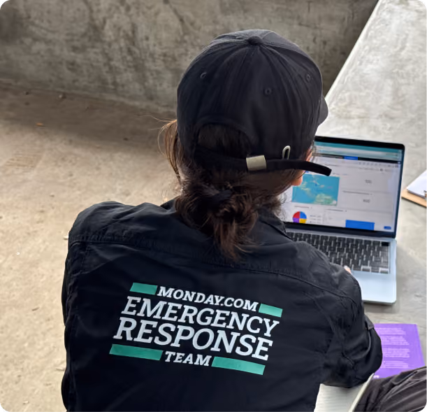 Person wearing a black cap and black jacket with 'Monday.com Emergency Response Team' printed on the back, working on a laptop showing data and charts. This image was taken from Jamaica hurricane on 2025.