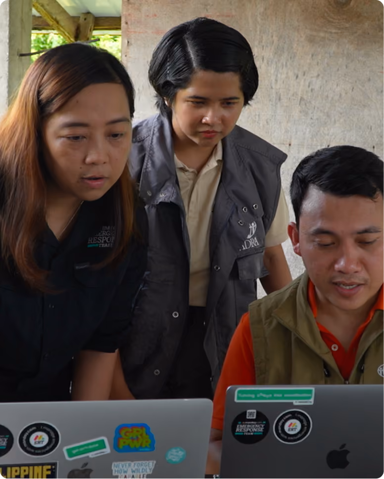 Three people closely looking at two laptops with various stickers, indoors near a window with wooden paneling in the background. The image is taken from Phillipines cyclone on 2025.