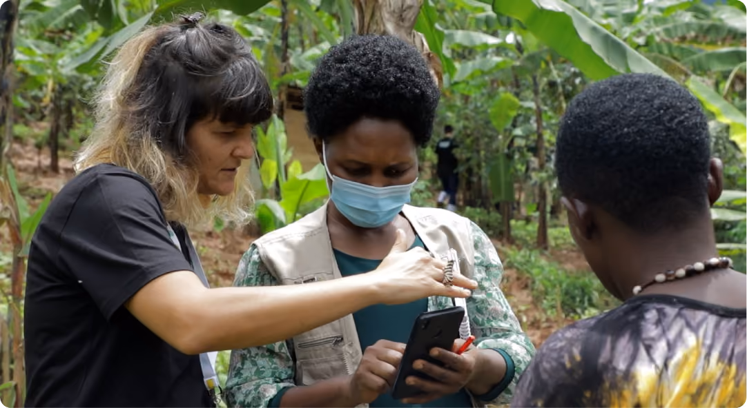 Three people in an outdoor setting with green foliage, one woman pointing at a smartphone held by another woman wearing a face mask. The image is taken from Uganda floods on 2022.