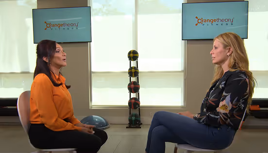 Two women seated facing each other in a fitness studio with Orangetheory Fitness logos on screens behind them.
