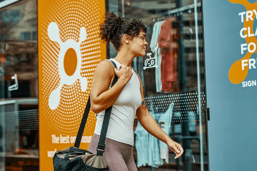 Woman in workout clothes carrying a gym bag walking past a fitness studio entrance with orange logos.