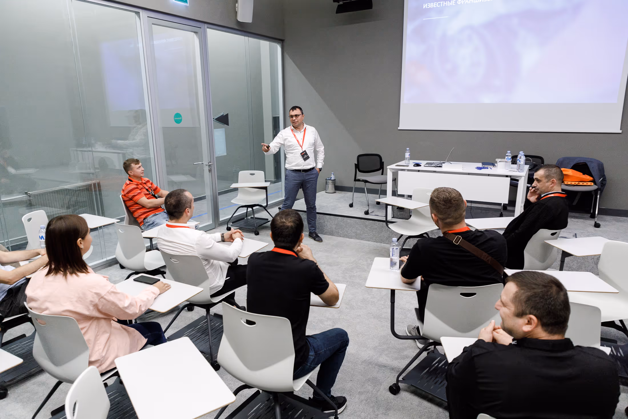 Мужчина с указкой ведёт презентацию перед группой слушателей в современном классе с проектором и стеклянными стенами.