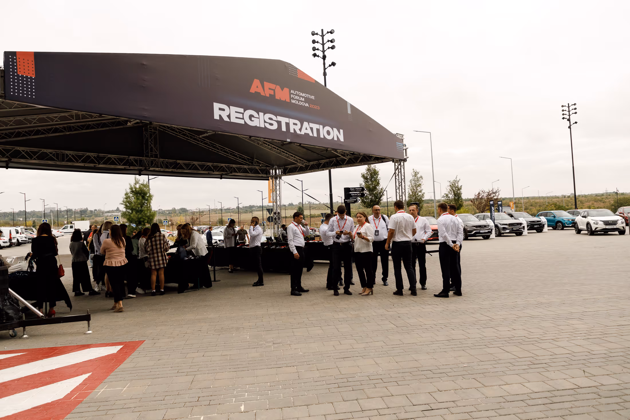 Группа людей с красными бейджами стоит и общается под шатром с надписью «Регистрация» на фоне припаркованных автомобилей и открытой местности.