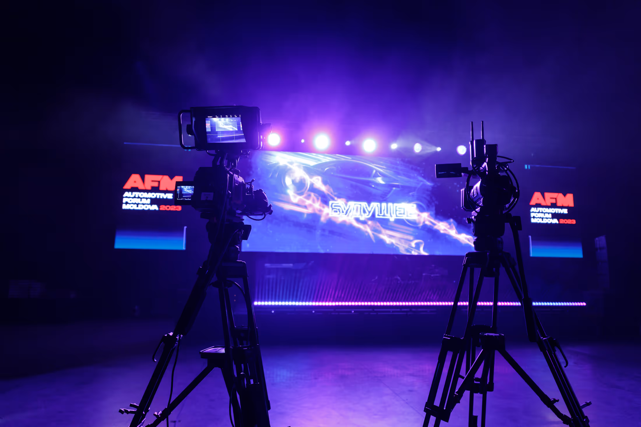 Два видеокамеры на штативах снимают сцену с большим экраном и надписью «Будущее» на форуме Automotive Forum Moldova 2023.