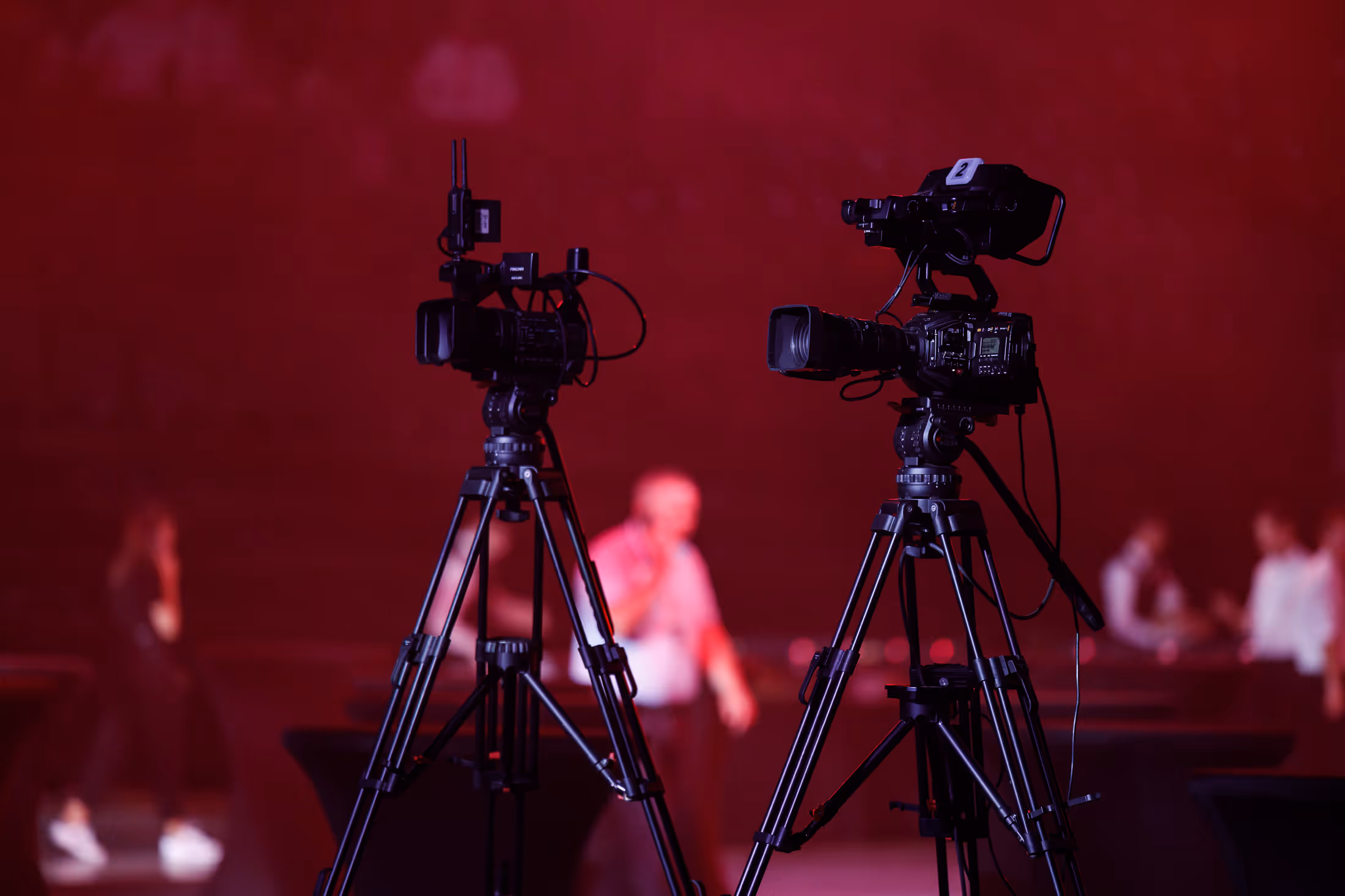 Две видеокамеры на штативах на фоне размытой сцены с людьми и красным освещением.