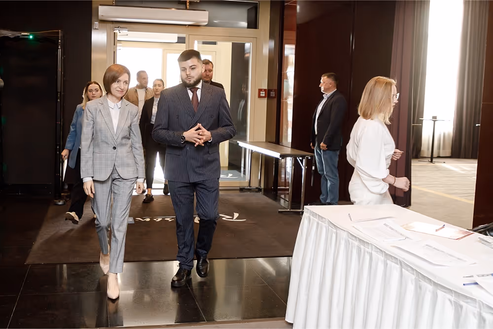 Группа людей в деловой одежде, включая мужчину и женщину в переднем плане, входят через дверной проем в светлое помещение.