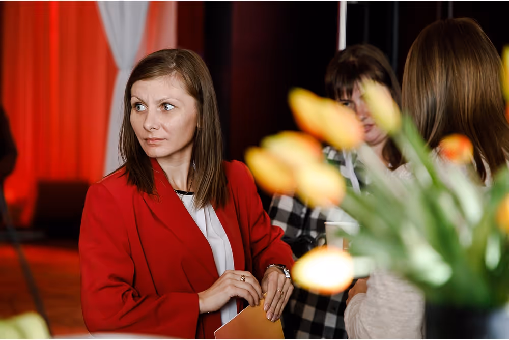 Женщина в красном пиджаке держит папку, на фоне размытые желтые цветы и две женщины, беседующие между собой.