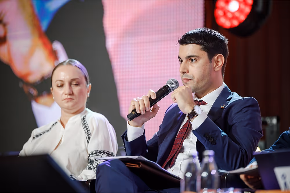 Мужчина в деловом костюме говорит в микрофон, женщина в вышиванке слушает на фоне сцены с ярким экраном.