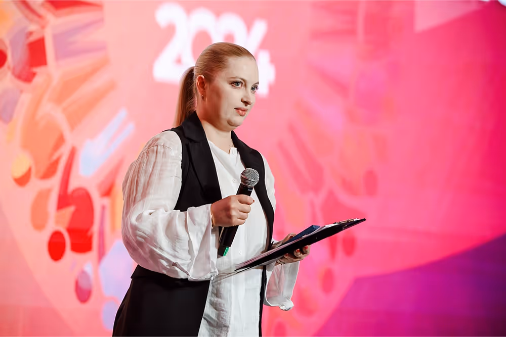 Женщина с микрофоном и планшетом выступает на фоне ярко-розового экрана с частично видимым текстом.