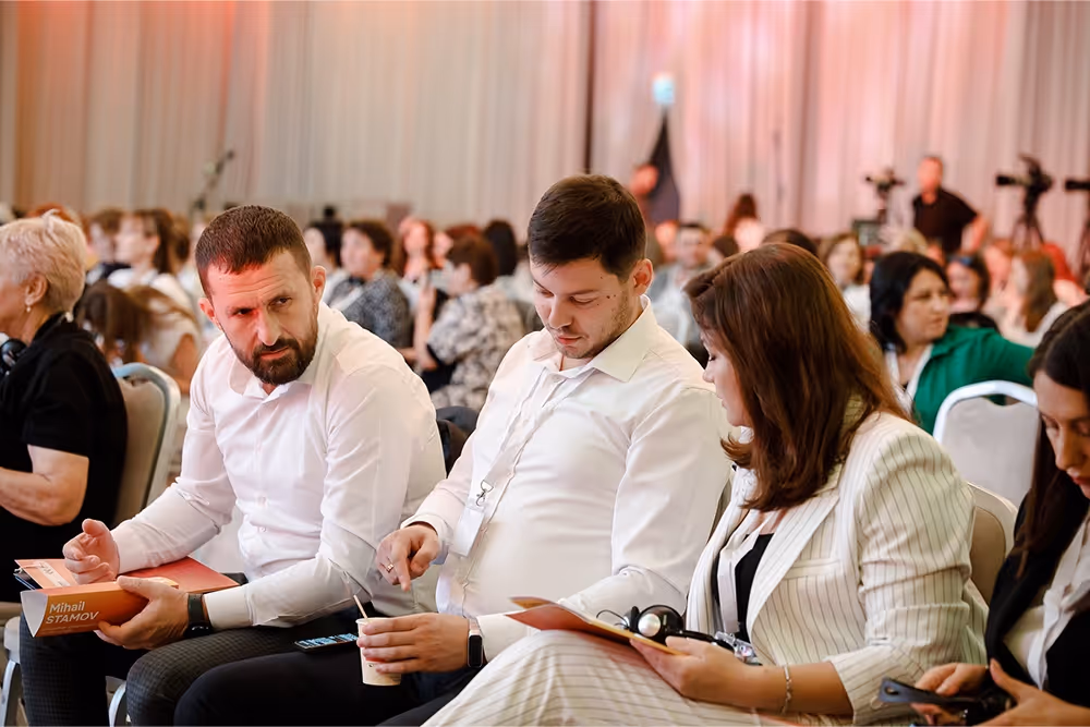 Группа взрослых людей, сидящих в конференц-зале, с тремя мужчинами и женщиной на переднем плане, обсуждающими материалы мероприятия.