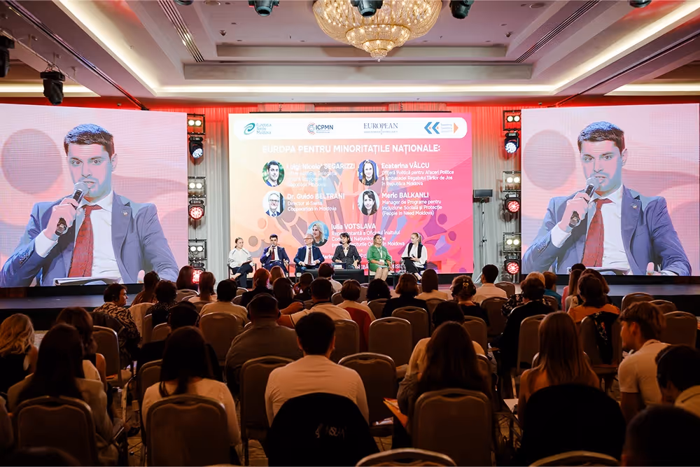 Конференция с панелистами на сцене и большим экраном с информацией о мероприятии на фоне, перед аудиторией.