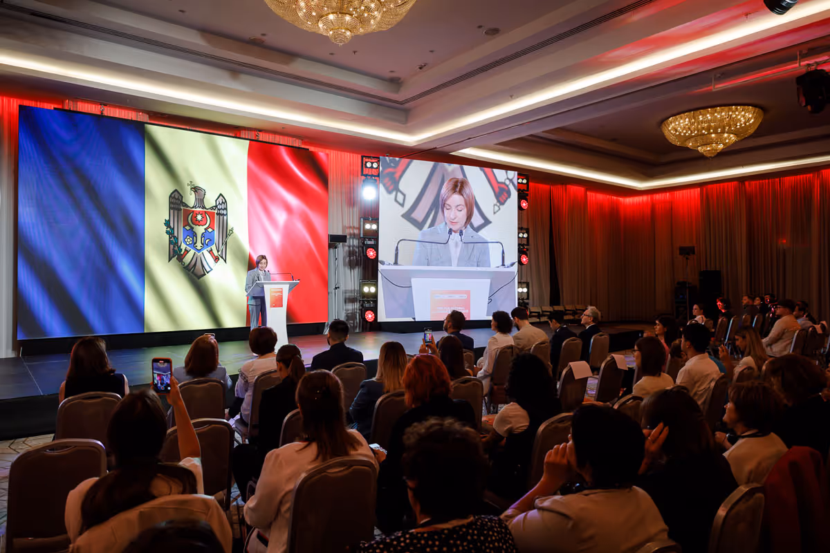 Женщина выступает на сцене перед большой аудиторией, на экране позади нее изображен молдавский флаг и ее увеличенное изображение.