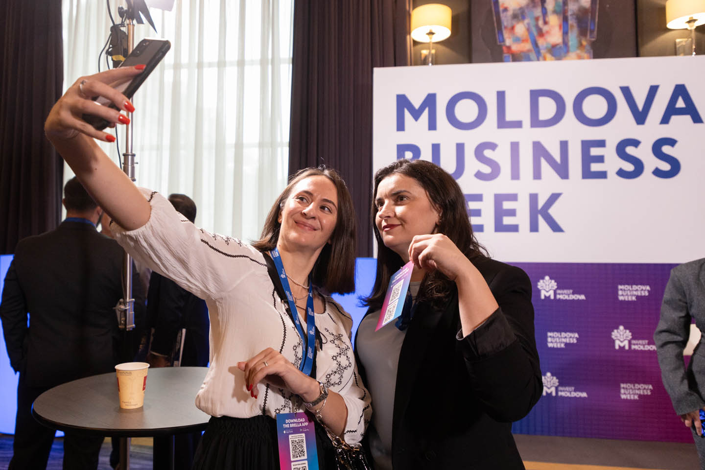 Две женщины делают селфи на фоне баннера Moldova Business Week, одна держит бейдж с QR-кодом.