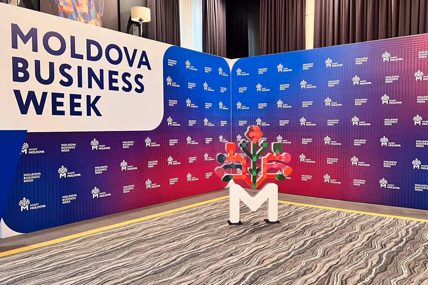 Стенд мероприятия Moldova Business Week с логотипом в форме буквы М и стилизованным графическим элементом.