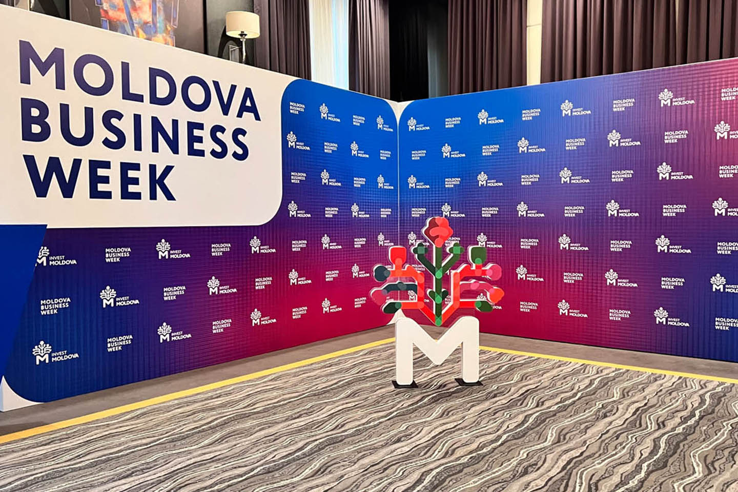 Стенд мероприятия Moldova Business Week с логотипом в форме буквы М и стилизованным графическим элементом.