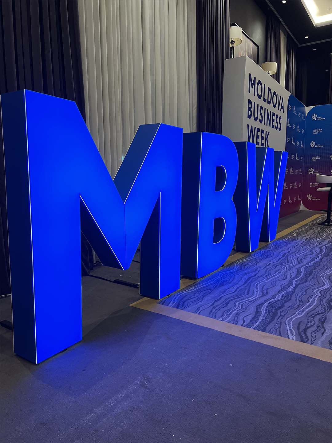 Большие светящиеся синие буквы MBW на полу в комнате с ковровым покрытием и занавесками, фон с надписью Moldova Business Week.
