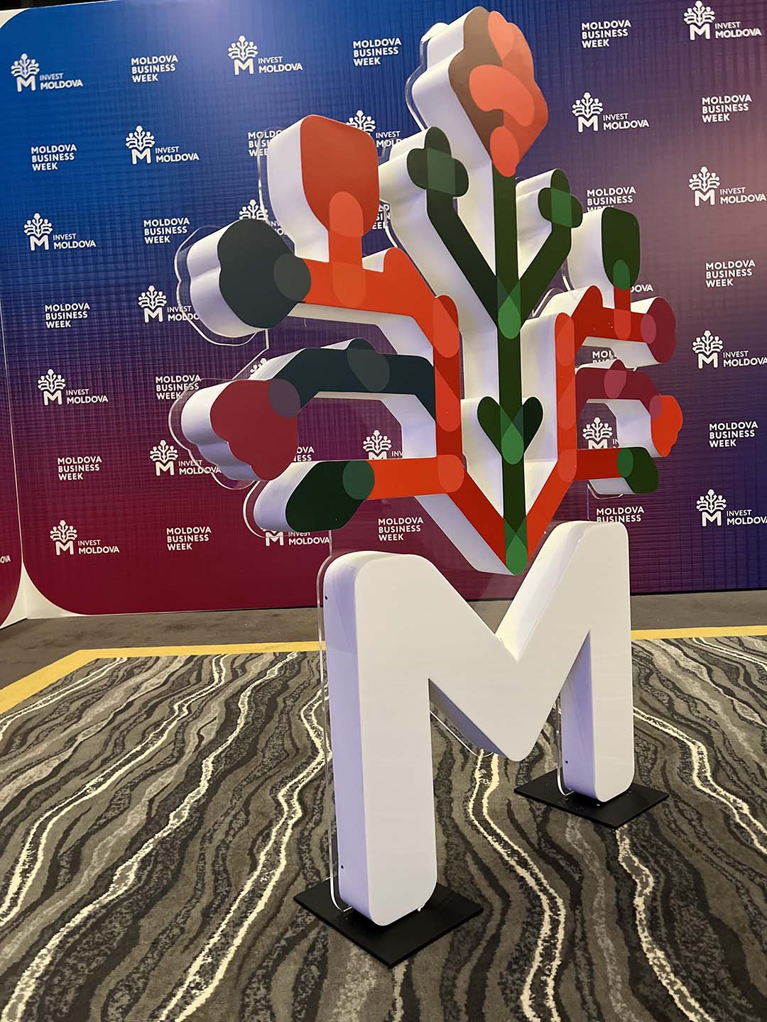 Большая белая буква М с цветочным логотипом Invest Moldova на фоне баннера Moldova Business Week.