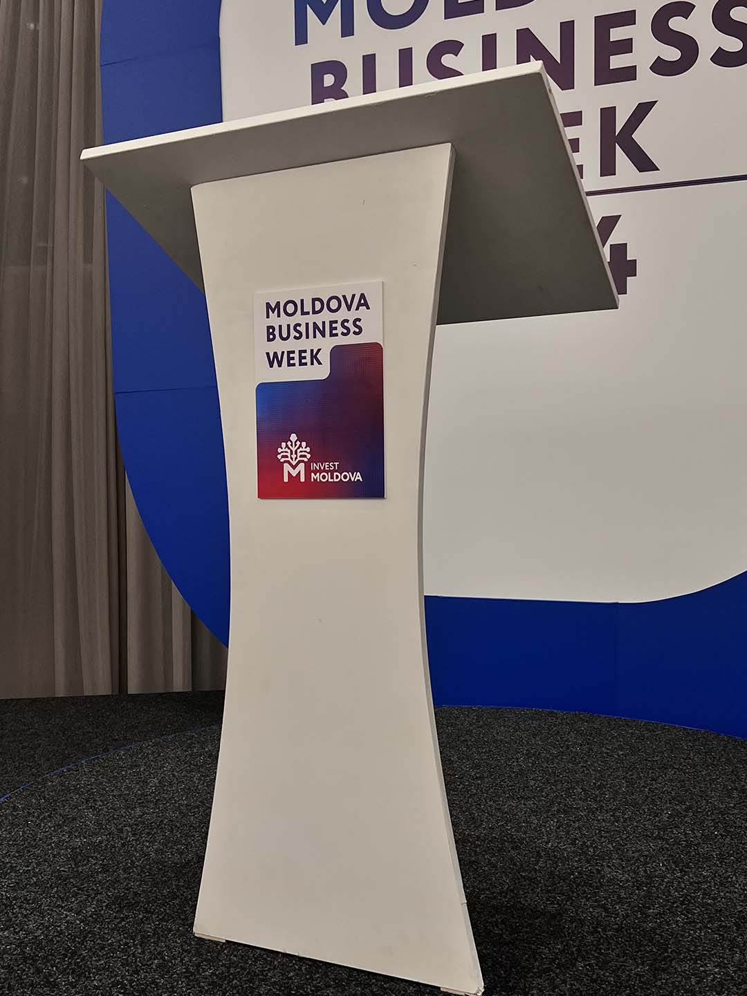 Белая трибуна с логотипом Moldova Business Week на фоне синего и белого баннера.