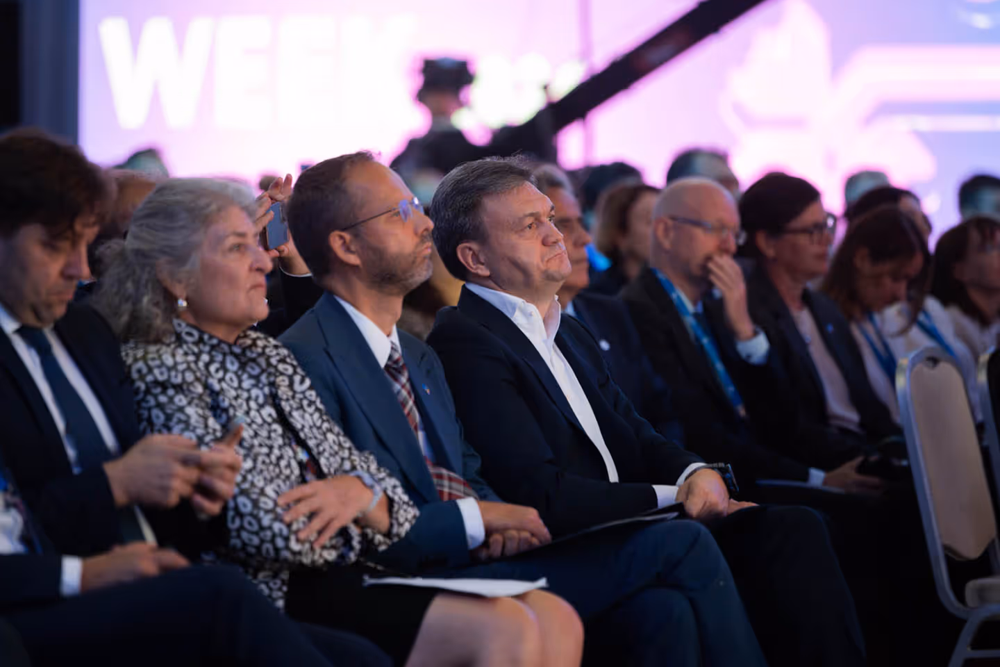 Группа взрослых людей в деловой одежде внимательно слушают презентацию на конференции.