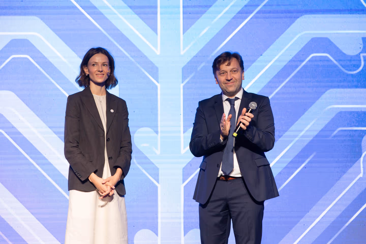 Мужчина в деловом костюме с микрофоном и женщина в светлом платье и темном пиджаке стоят на сцене на фоне синего абстрактного узора.