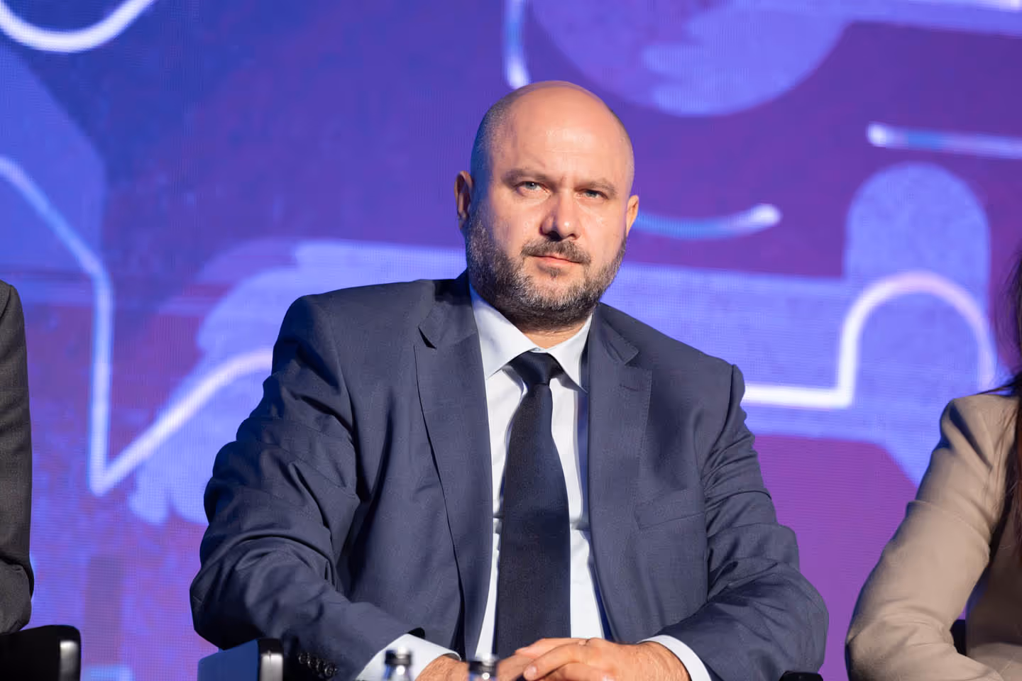 Мужчина в деловом костюме и галстуке сидит на конференции на фоне пурпурного экрана.