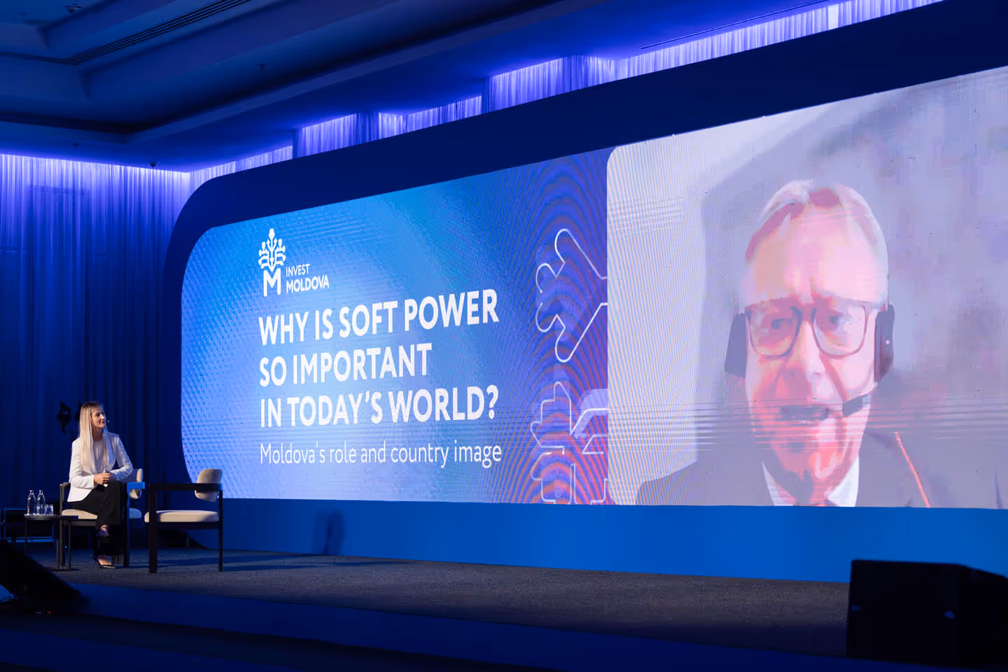 Женщина на сцене смотрит на большой экран с надписью «Почему мягкая сила так важна в современном мире? Роль Молдовы и имидж страны», на экране изображен мужчина с гарнитурой.