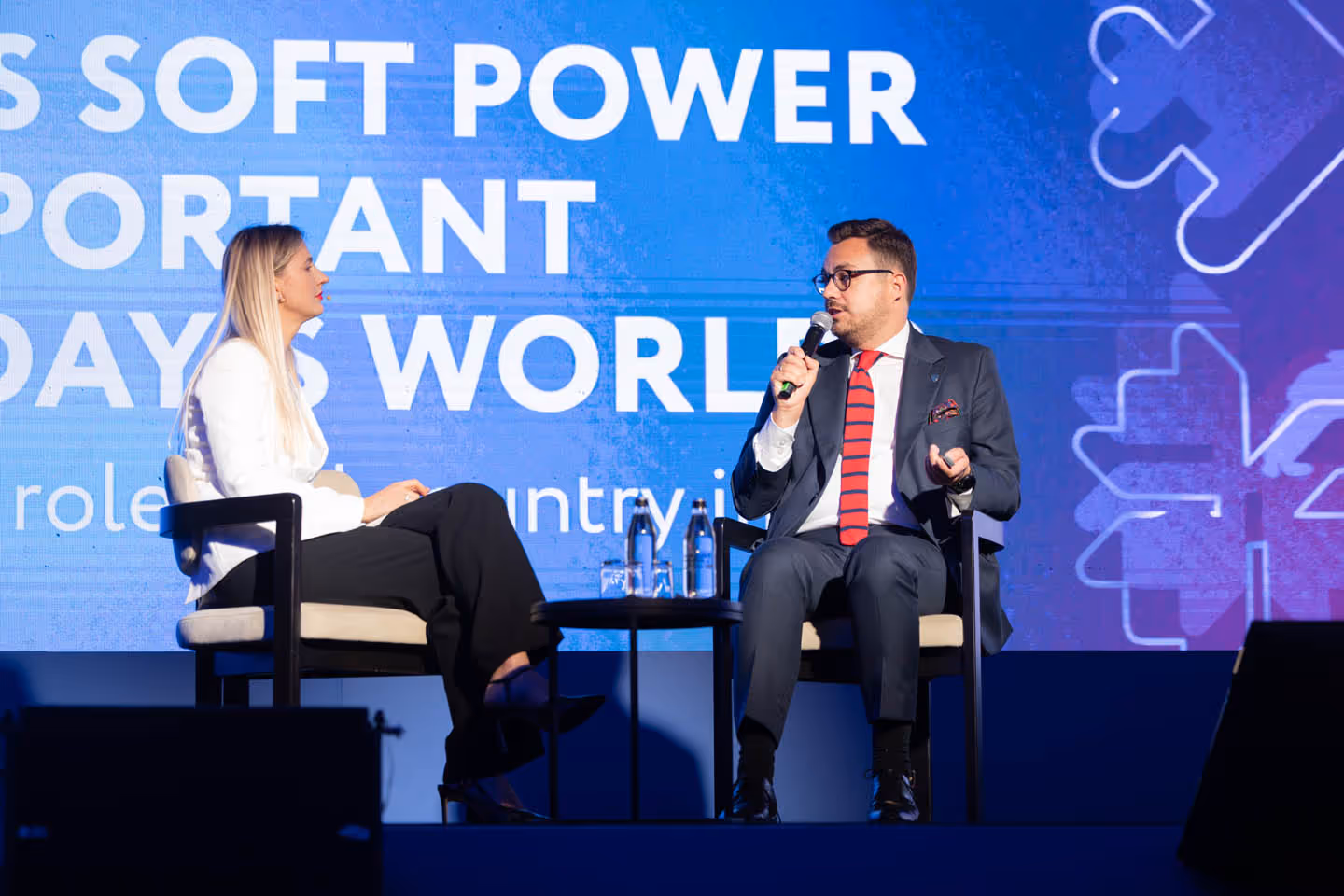 Два человека, мужчина и женщина, сидят на стульях на сцене и ведут беседу в микрофон на фоне большого экрана с текстом.