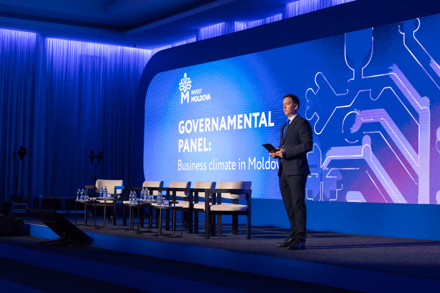 Мужчина в костюме стоит на сцене у большого экрана с надписью "GOVERNAMENTAL PANEL: Business climate in Moldova", рядом пустые стулья и столы с бутылками воды.